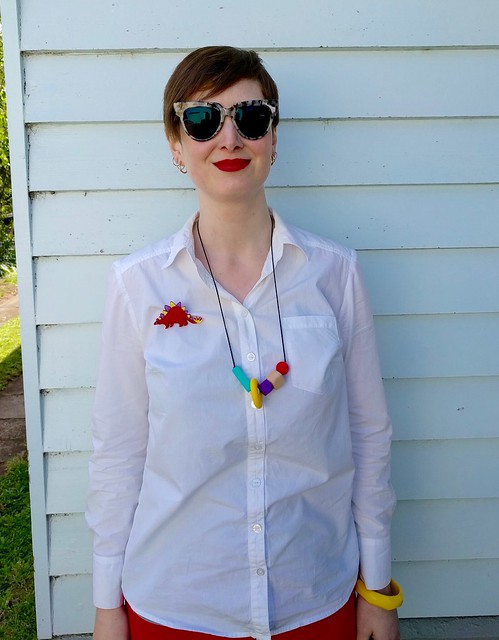 A woman wears a white button up shirt, colourful necklace and dinosaur brooch.