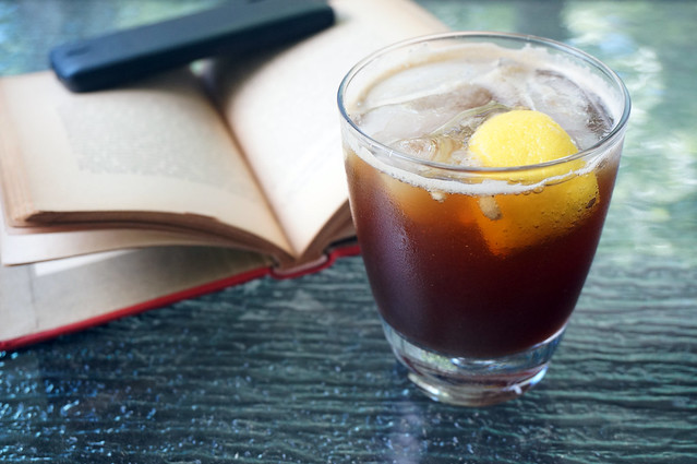 The drink sits on a glass table, dark brown and inviting. In the background, a book with age-yellowed pages is held open by a black smartphone.